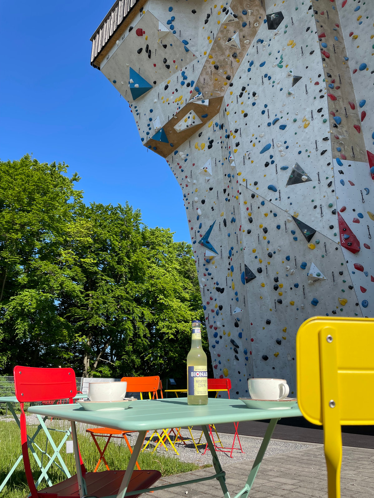Sonnige Terrasse des KletterZ’ mit bunten Stühlen, Kaffeetassen und der hohen Außenkletterwand im Hintergrund.